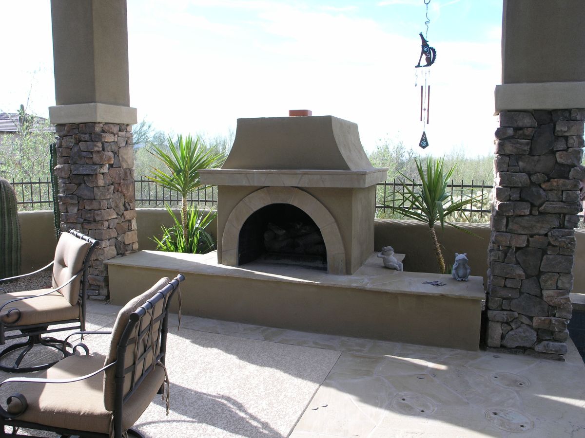 Outdoor patio fireplace with built-in stucco hearth, stacked-stone columns, cushioned iron chairs, desert plants