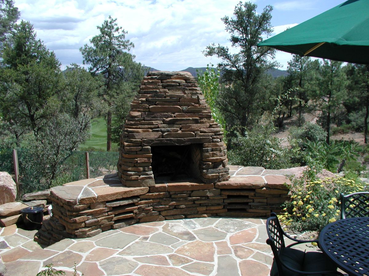 Outdoor stone fireplace on flagstone patio with curved stone bench, garden seating and green umbrella in wooded backyard