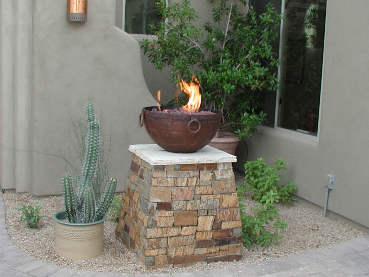 Outdoor rustic fire bowl on stacked-stone pedestal in desert courtyard with potted cactus and stucco wall