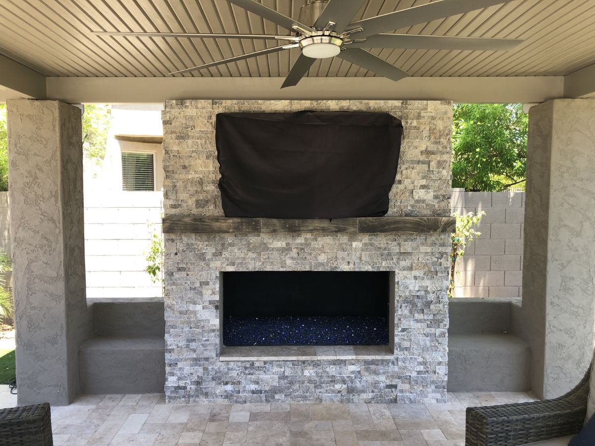 Outdoor covered patio with stone veneer fireplace, wood mantel, blue fire-glass bed, large ceiling fan and covered TV