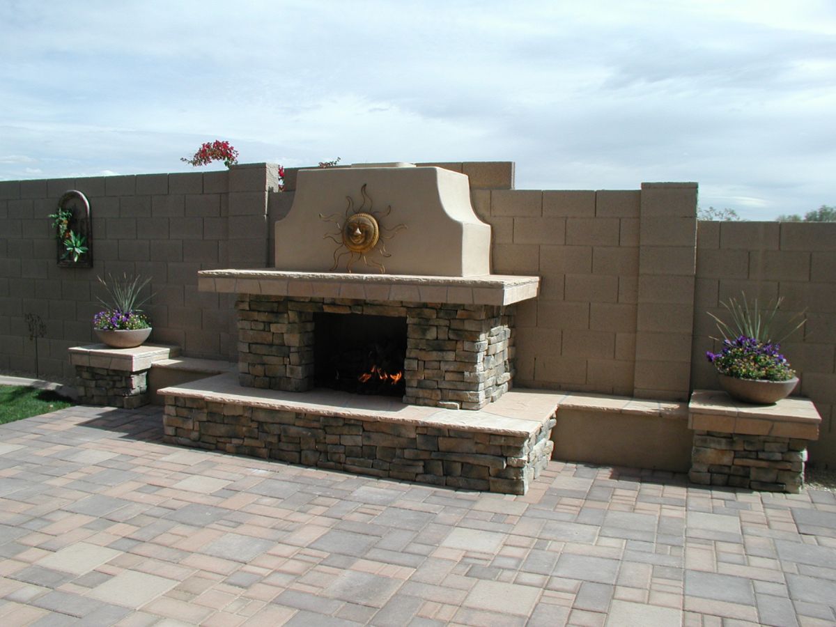 Outdoor stone fireplace on paver patio with sun wall decor and potted plants, backyard seating area