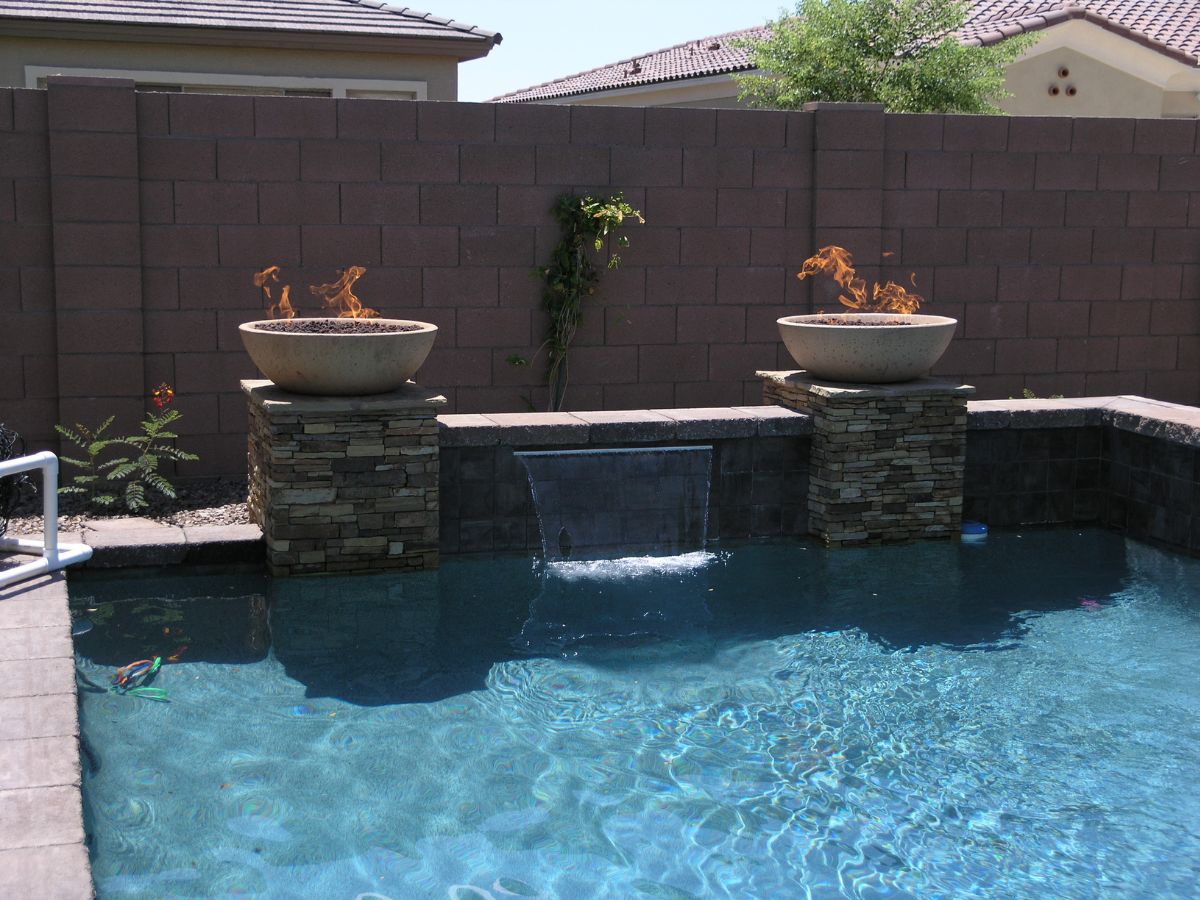 Backyard pool with clear blue water, central waterfall and two fire bowls on stacked-stone pillars.