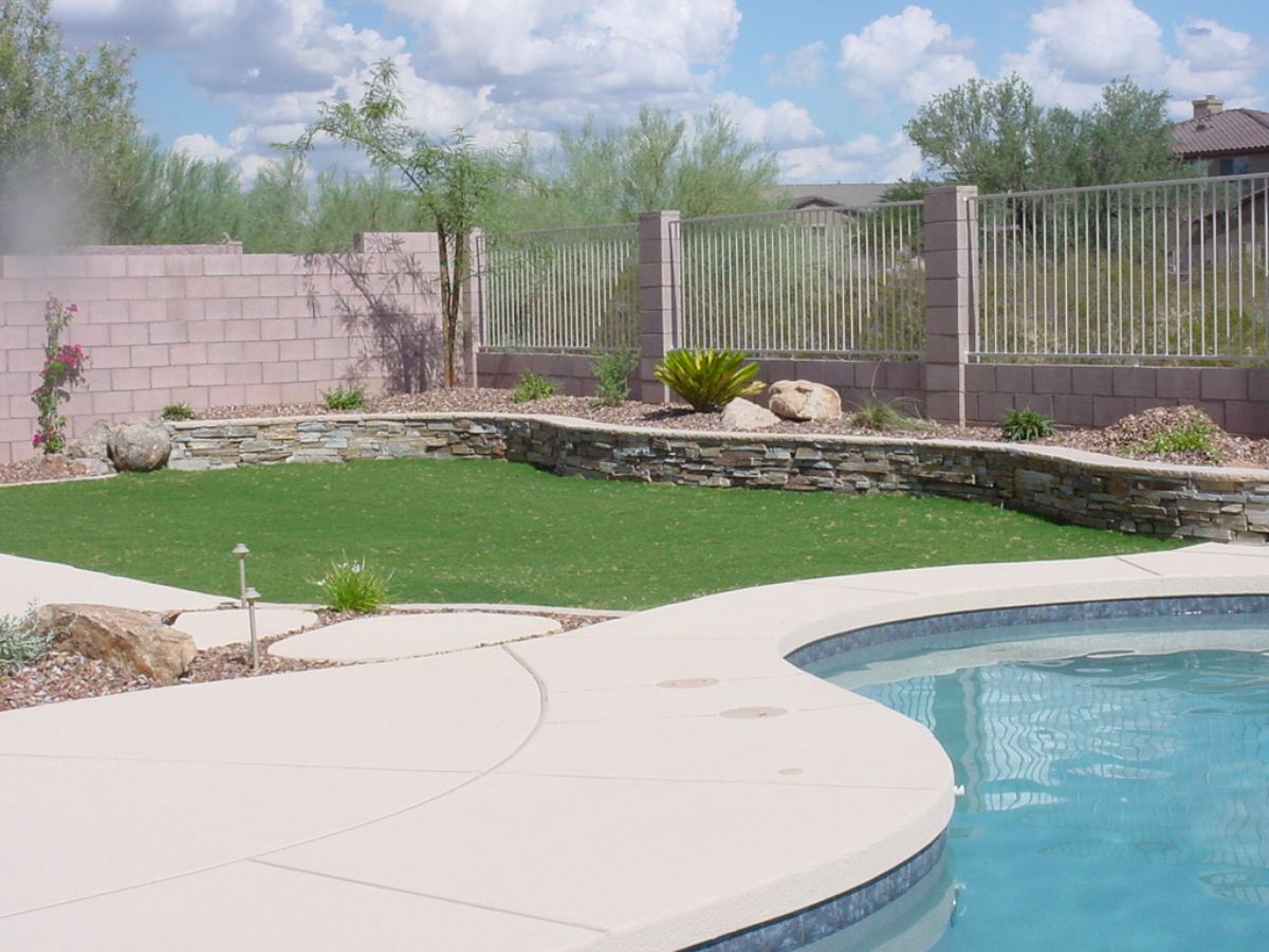 Backyard swimming pool with curved concrete patio, artificial turf, stone retaining wall, desert landscaping