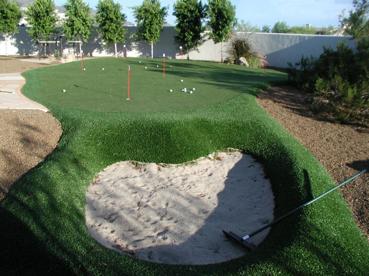Backyard artificial putting green with sand bunker, rake, red flagsticks and scattered golf balls for practice