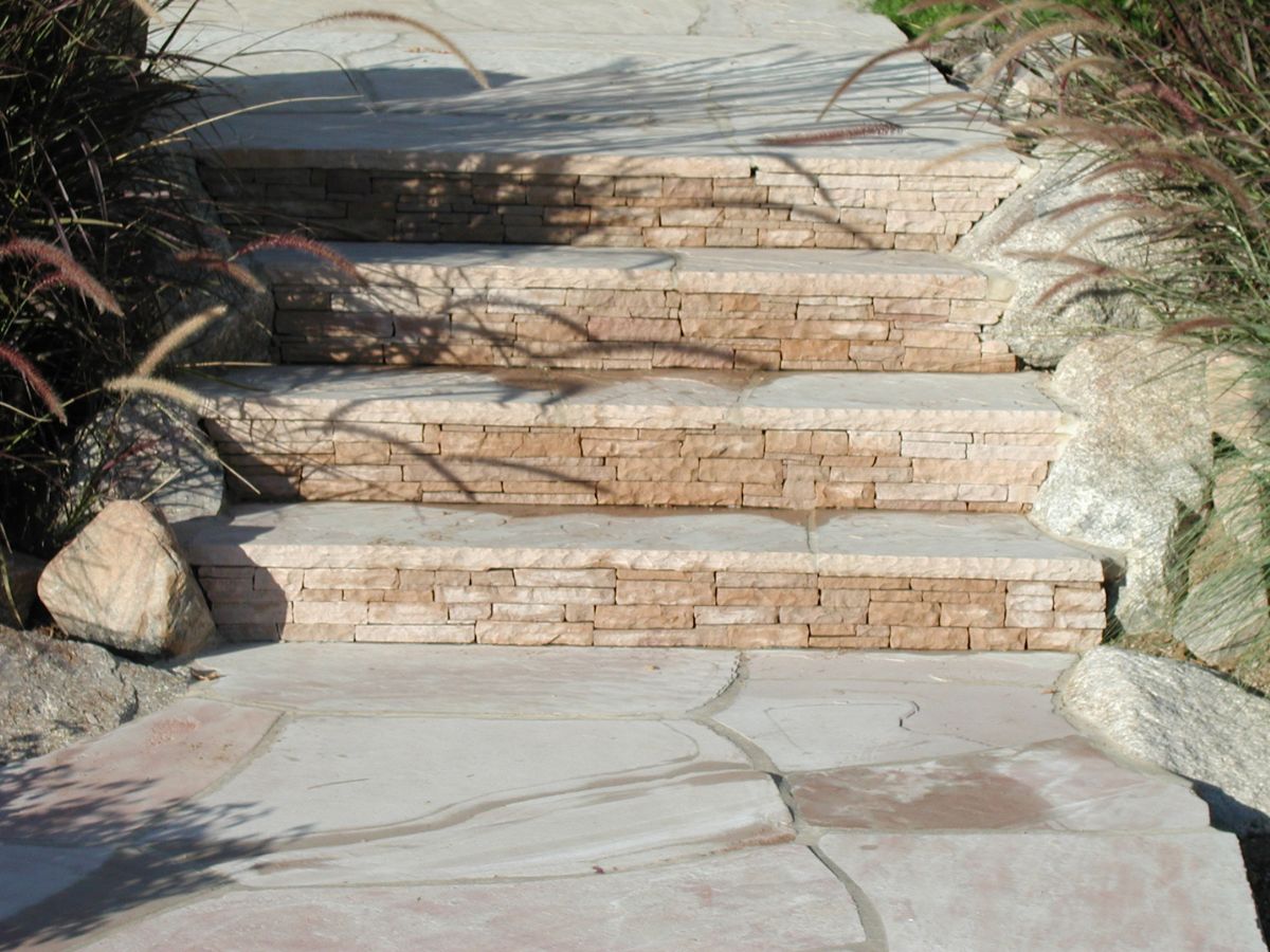 Sunlit stacked stone steps and flagstone walkway flanked by ornamental grasses and boulders in a landscaped garden