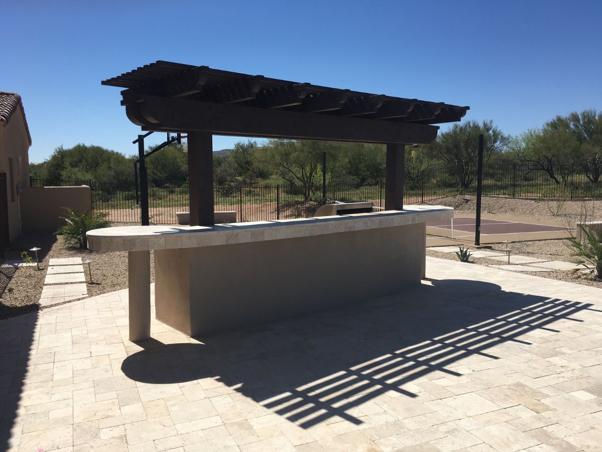 Desert backyard patio with pergola-covered outdoor stone bar, tiled paver flooring and fenced sports court beyond.