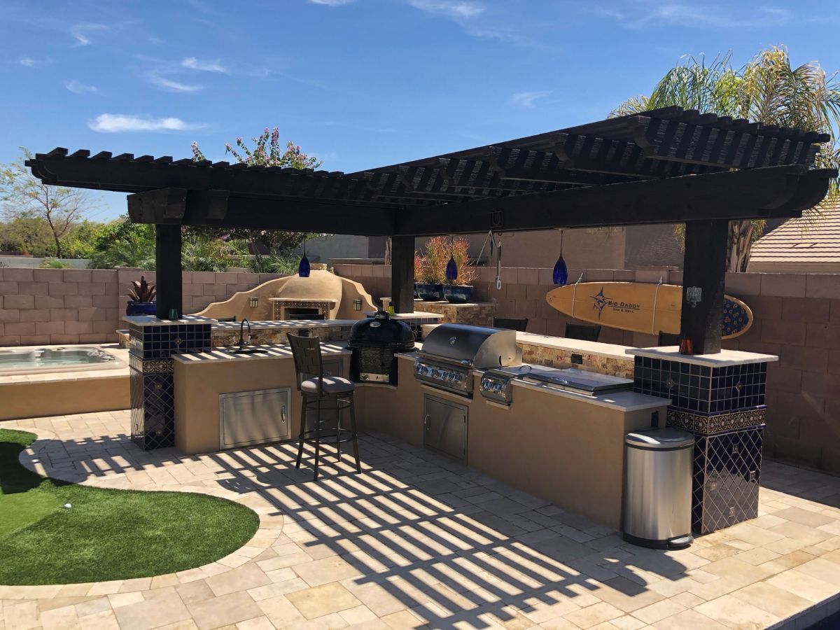 Outdoor kitchen pergola with built-in stainless grill, bar seating, tile columns and hot tub on sunlit patio