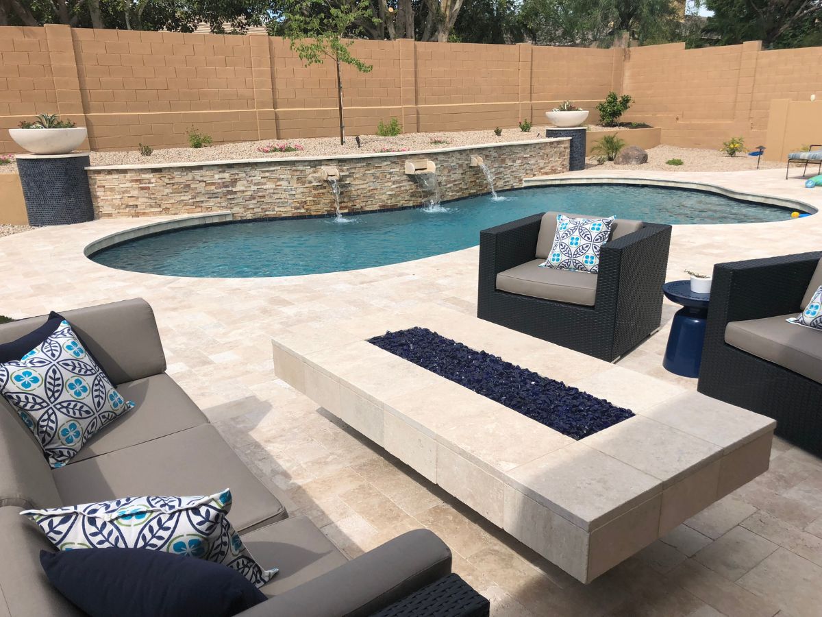 Modern backyard pool and patio with travertine pavers, wicker seating, patterned pillows, and glass-filled linear fire pit.