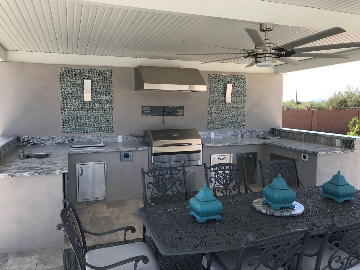 Covered outdoor kitchen with stainless grill, stone countertops, ceiling fan, and wrought-iron dining table with teal accents