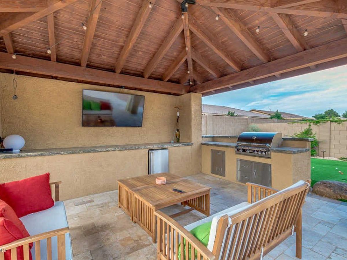Covered patio outdoor kitchen with wood-beamed ceiling, built-in stainless grill, wall TV, wooden seating and coffee table