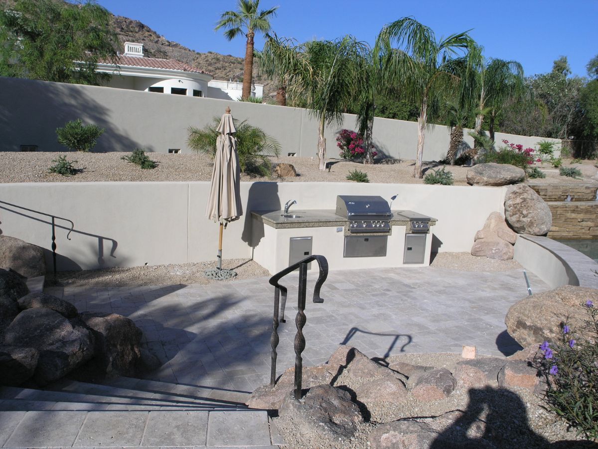 Modern outdoor kitchen with built-in stainless grill, sink and umbrella on tiled patio, desert landscaping and palm trees.