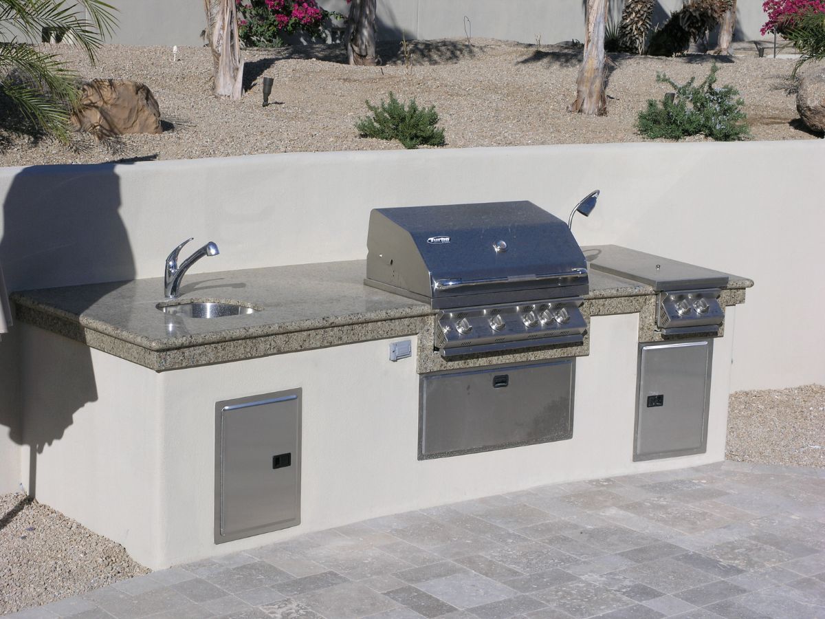 Outdoor kitchen with built-in stainless steel gas grill, side burner, granite countertop and sink on stone patio