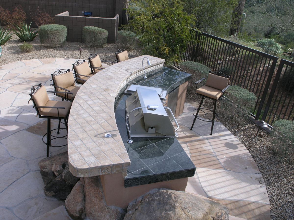 Outdoor patio kitchen with curved stone bar, built-in stainless grill and cushioned bar stools, desert landscaping