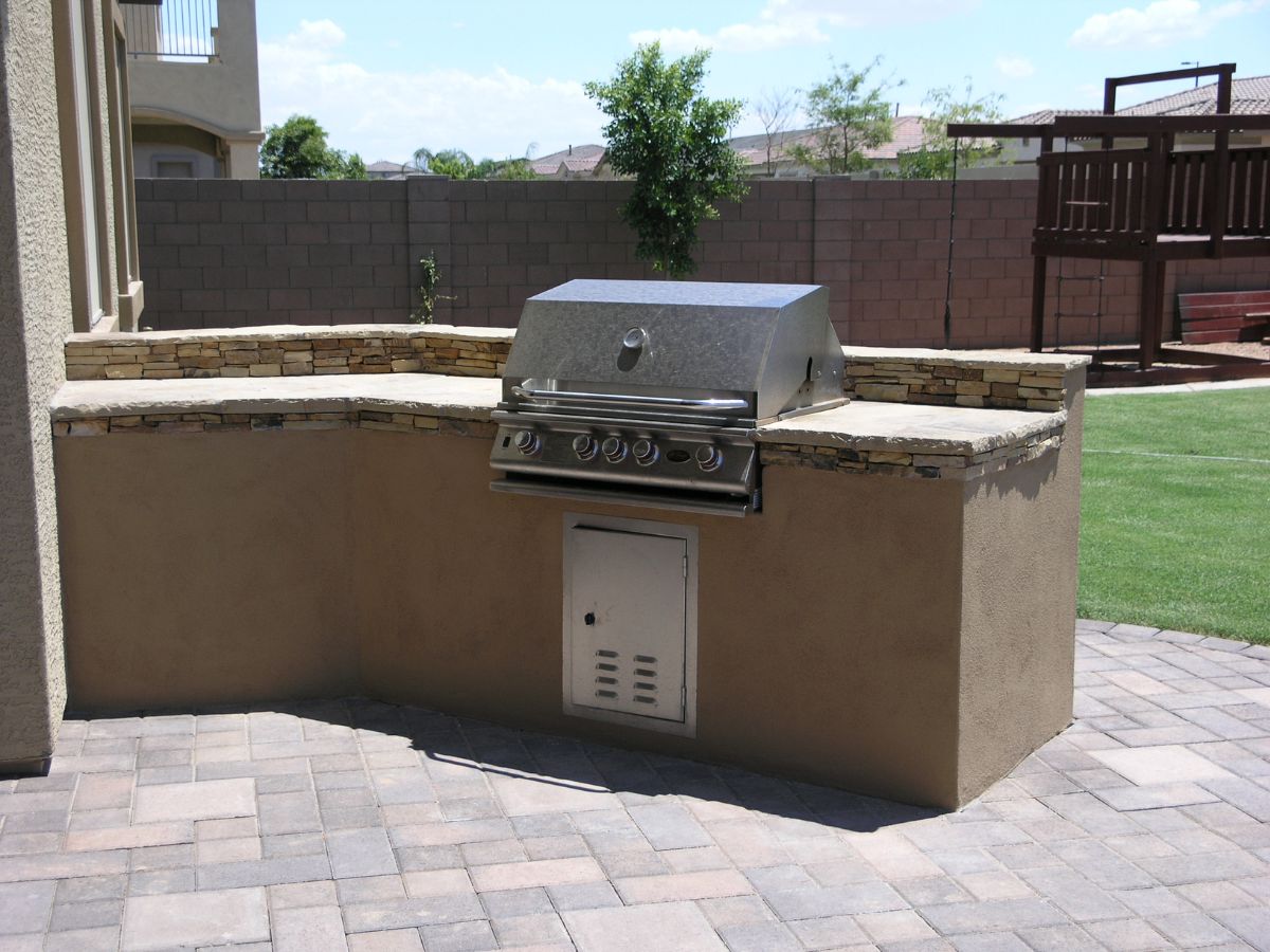Built-in stainless steel gas grill in stone counter on paver patio with green backyard lawn