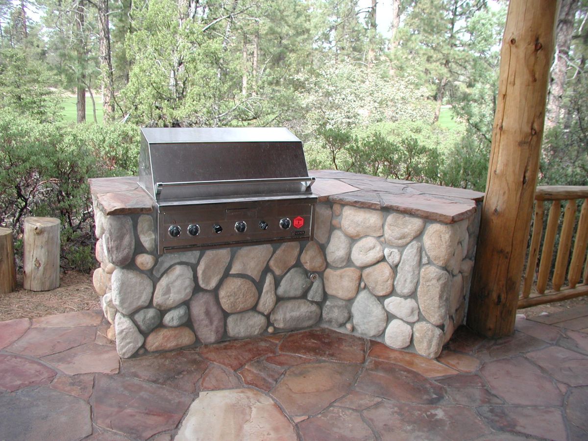 Stainless steel built-in outdoor grill in river rock stone island on rustic flagstone patio in wooded backyard