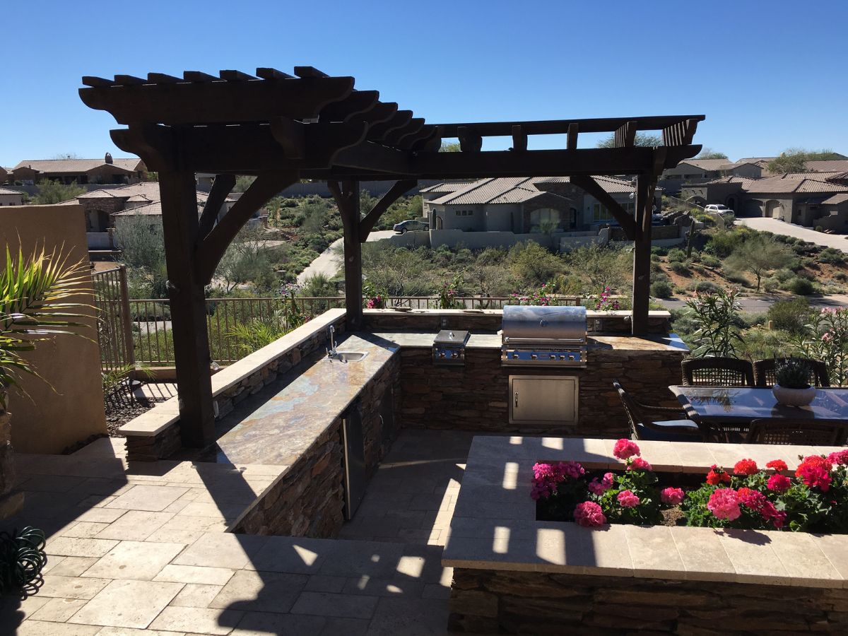 Outdoor kitchen and dining patio with wooden pergola, stone countertops, built-in stainless grill and desert landscape view.