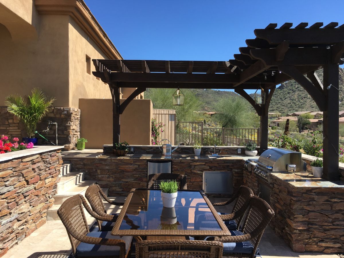 Sunlit backyard outdoor kitchen with wooden pergola, stone countertops, gas grill and wicker dining set with mountain view