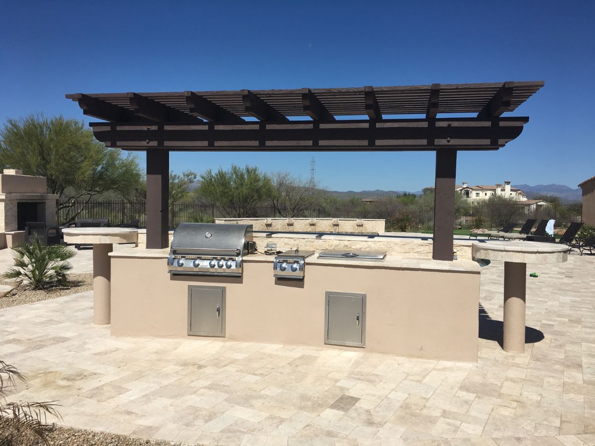 Poolside outdoor kitchen with stainless steel built-in grill, stone countertop and wooden pergola in desert backyard.