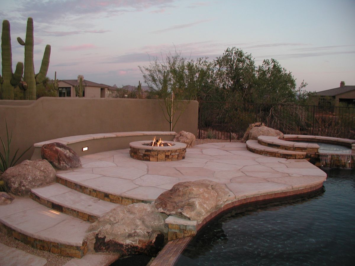 Desert backyard patio with stone decking, circular fire pit, steps and pool, saguaro cacti and evening sky
