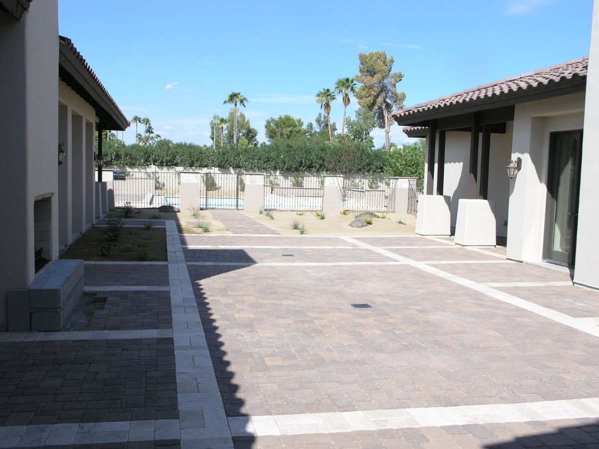 Sunlit Southwestern villa courtyard with patterned paver patio, covered walkways, palm trees and gated pool beyond.