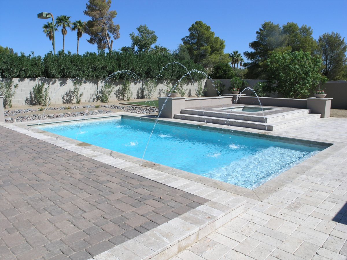 Modern backyard swimming pool with arcing fountain jets, raised spa, and stone paver patio under blue sky