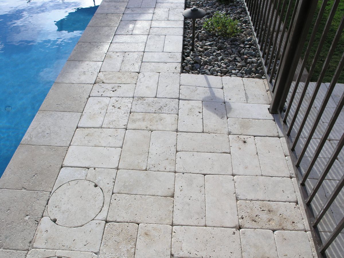 Travertine pool deck pavers and coping next to blue swimming pool, metal safety fence and decorative river rock bed