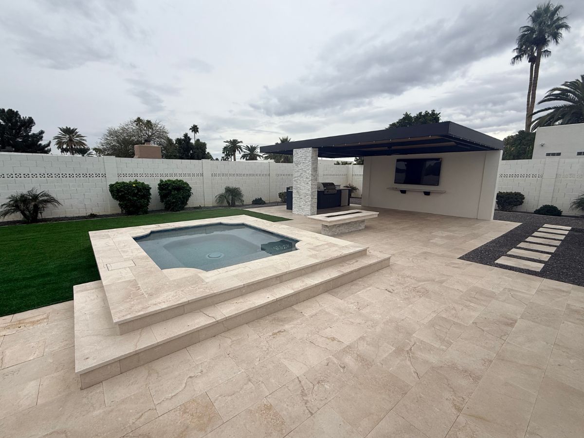 Backyard patio with raised travertine spa, covered outdoor kitchen, wall-mounted TV, palm trees and manicured lawn.