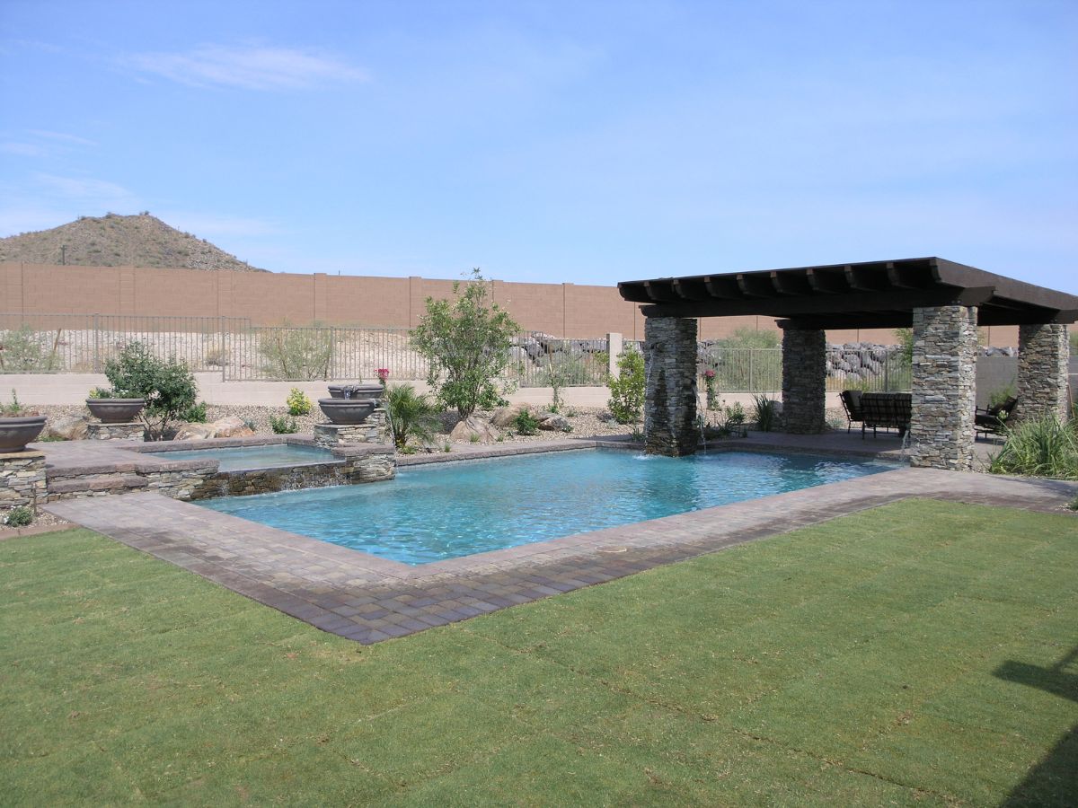 Luxury backyard swimming pool with stacked-stone pergola, paved deck, manicured lawn and desert mountain backdrop
