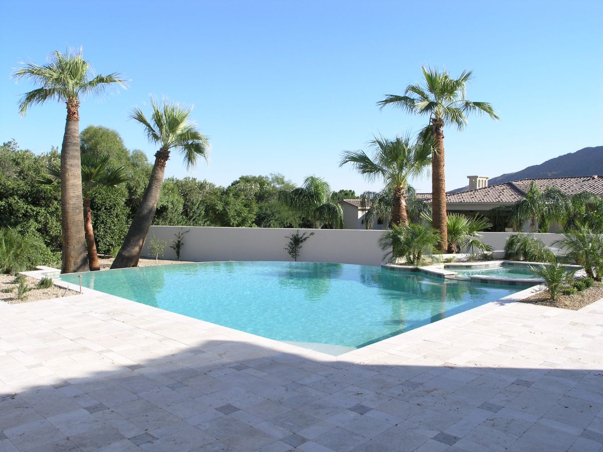 Luxury infinity pool with clear blue water, palm trees and travertine patio in upscale backyard by Mediterranean-style house