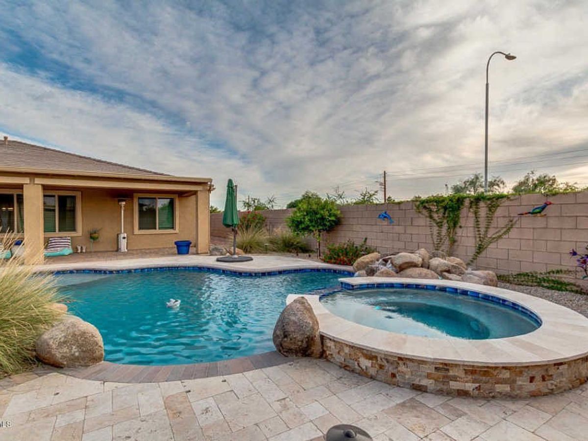 Backyard pool and spa with rock waterfall, tiled patio, umbrella and landscaping beside single-story home