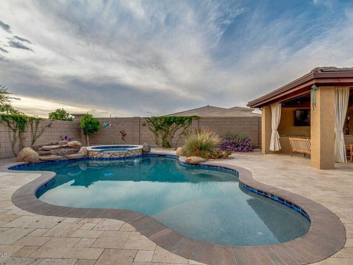 Kidney-shaped backyard pool with built-in spa, stone decking and covered patio with curtains