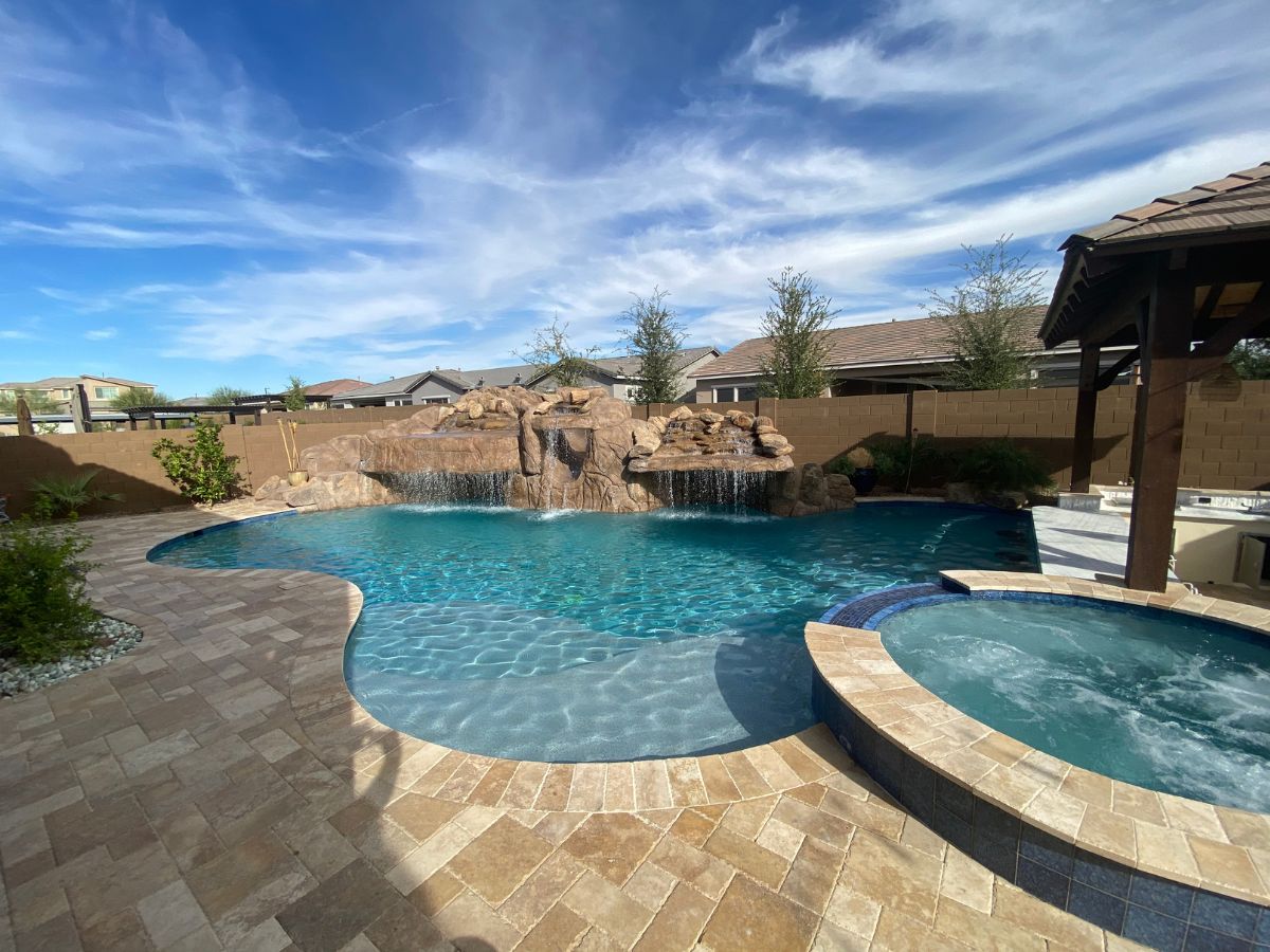 Luxury backyard swimming pool with rock waterfall, attached spa and travertine paver patio under blue sky