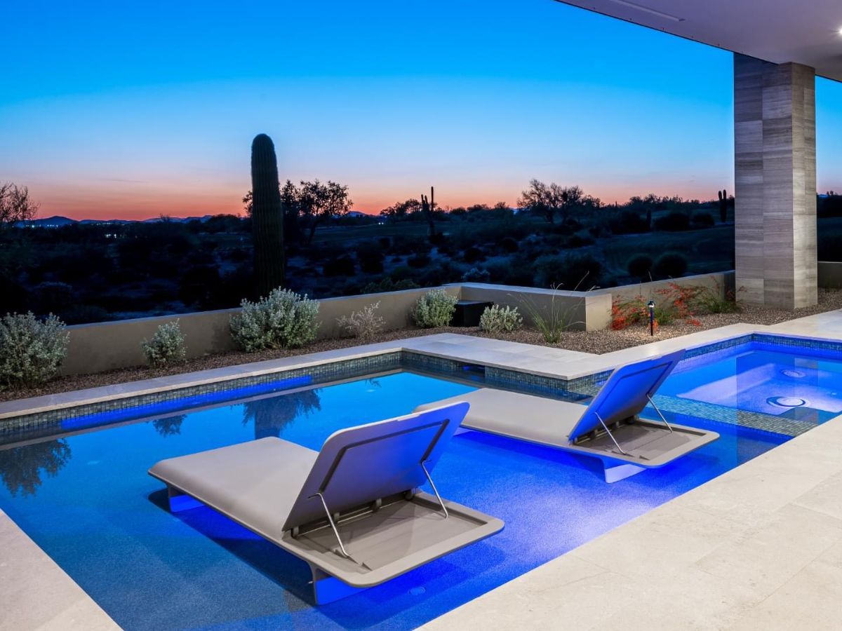 Modern luxury pool with illuminated blue water and two in-pool lounge chairs overlooking desert sunset with saguaro cactus
