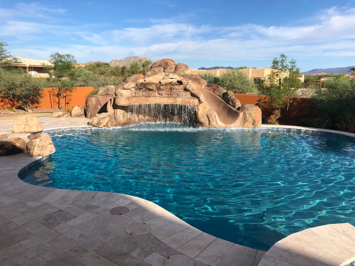 Luxury backyard swimming pool with rock waterfall and waterslide, blue water, travertine deck and desert landscaping