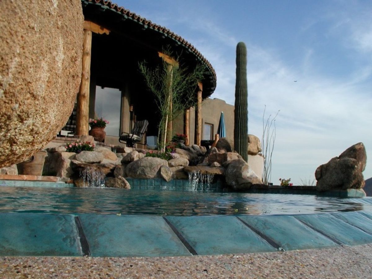 Turquoise infinity-edge pool with boulder waterfall beside a Southwestern desert villa and tall saguaro cactus under blue sky
