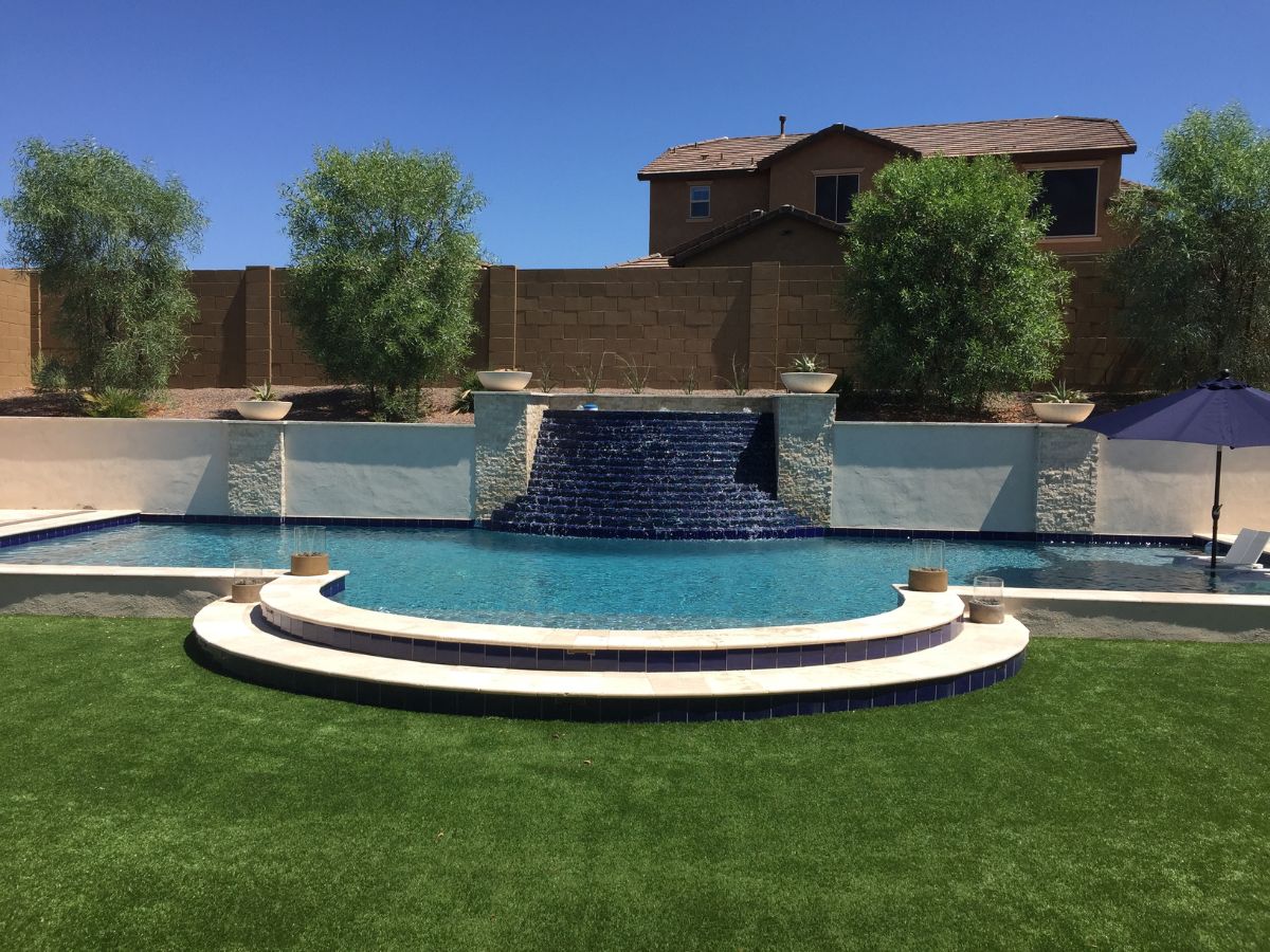 Luxury backyard pool with blue-tiled waterfall, curved steps, patio urns and artificial turf lawn under clear sky