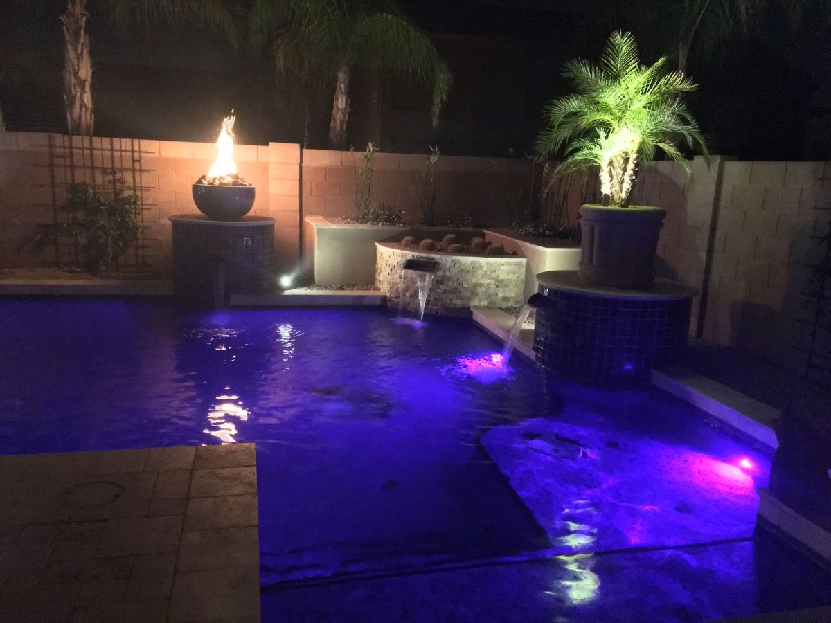 Luxury backyard pool at night with purple LED lights, cascading water features, flaming fire bowl and illuminated potted palm.