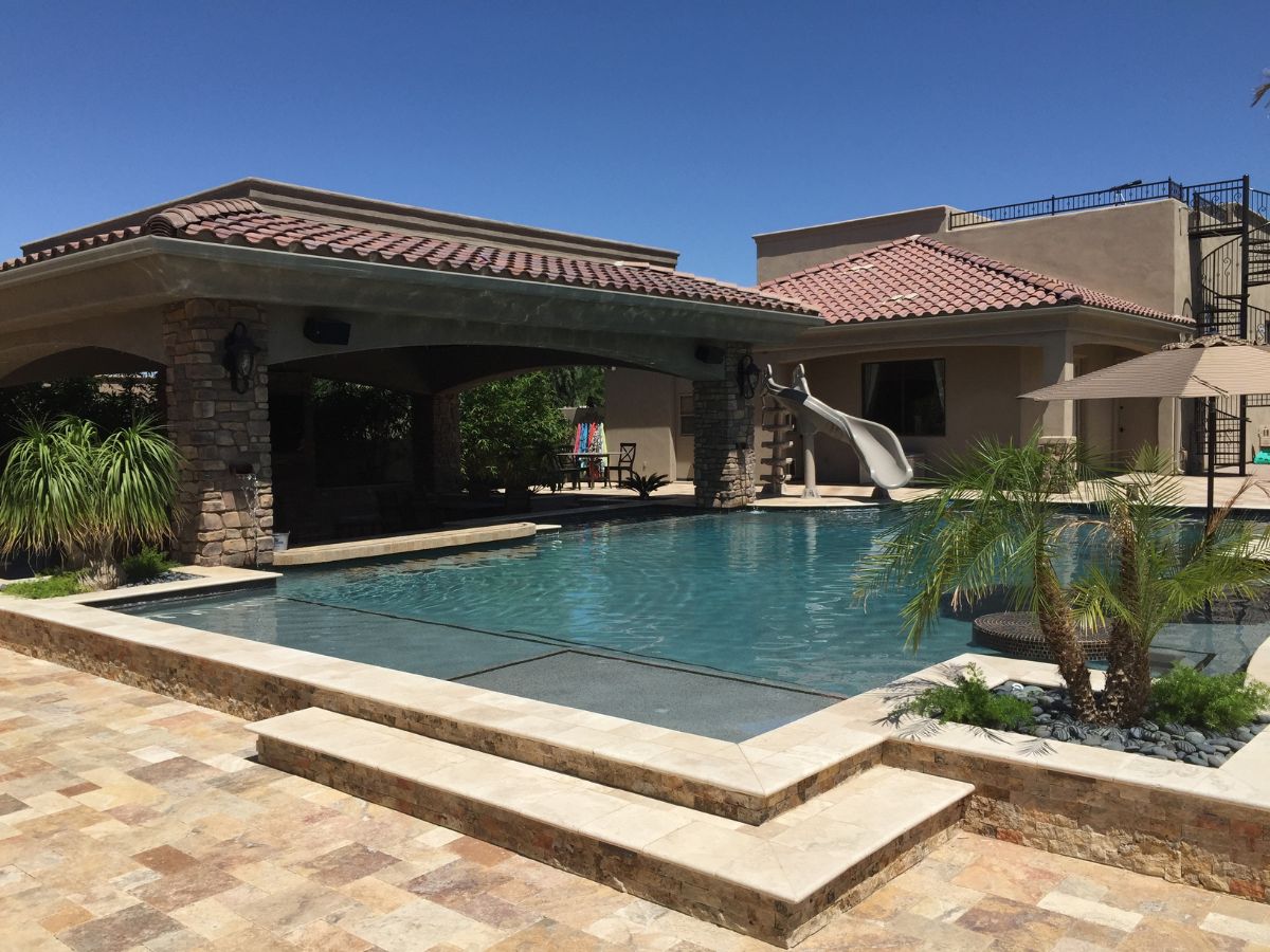 Luxury backyard pool with water slide, covered stone patio, palm landscaping and travertine tile deck at Mediterranean home