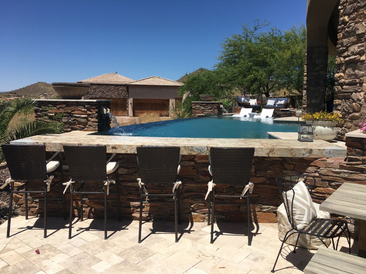 Resort-style backyard infinity pool with stone bar, four wicker bar stools and lounge seating under clear blue sky