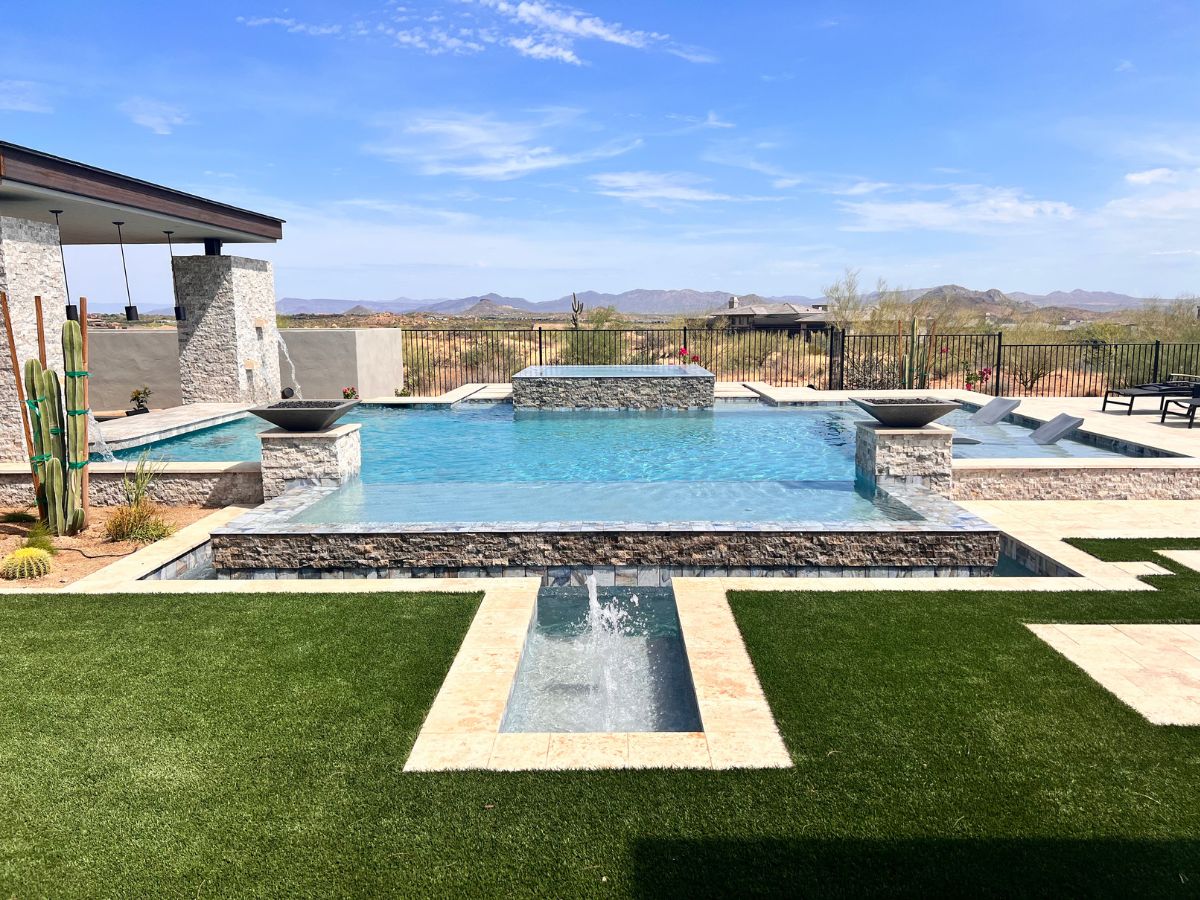 Luxury modern backyard infinity pool and spa with stone accents, sun loungers, artificial turf and desert mountain views.