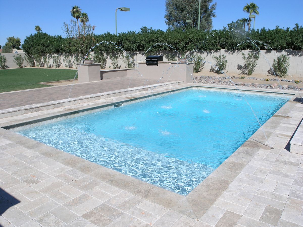 Rectangular backyard swimming pool with blue water, stone-paver patio and arcing water jets under clear sky