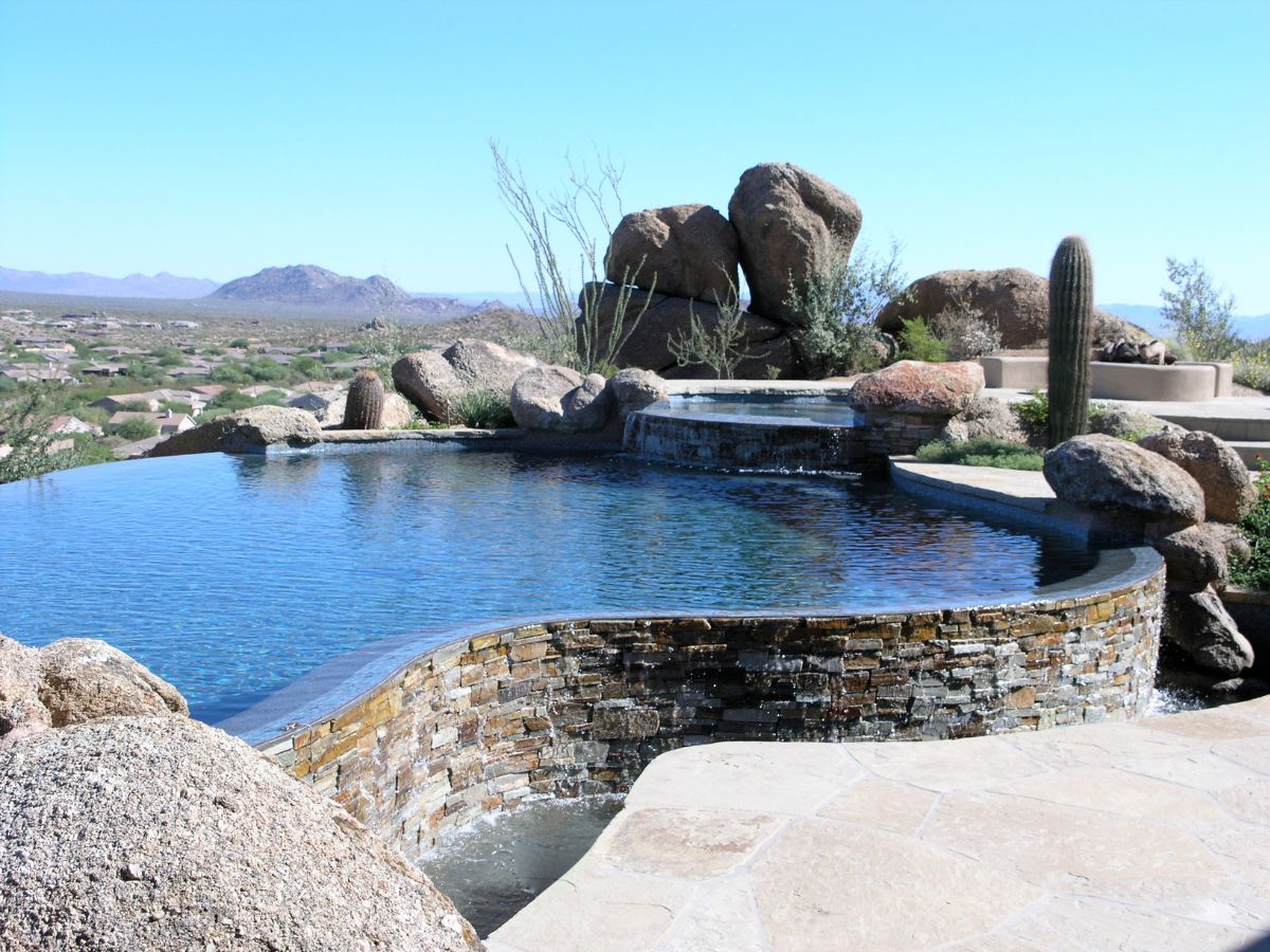 Infinity-edge luxury pool with stone waterfall, boulders and cactus overlooking a desert valley and distant mountains
