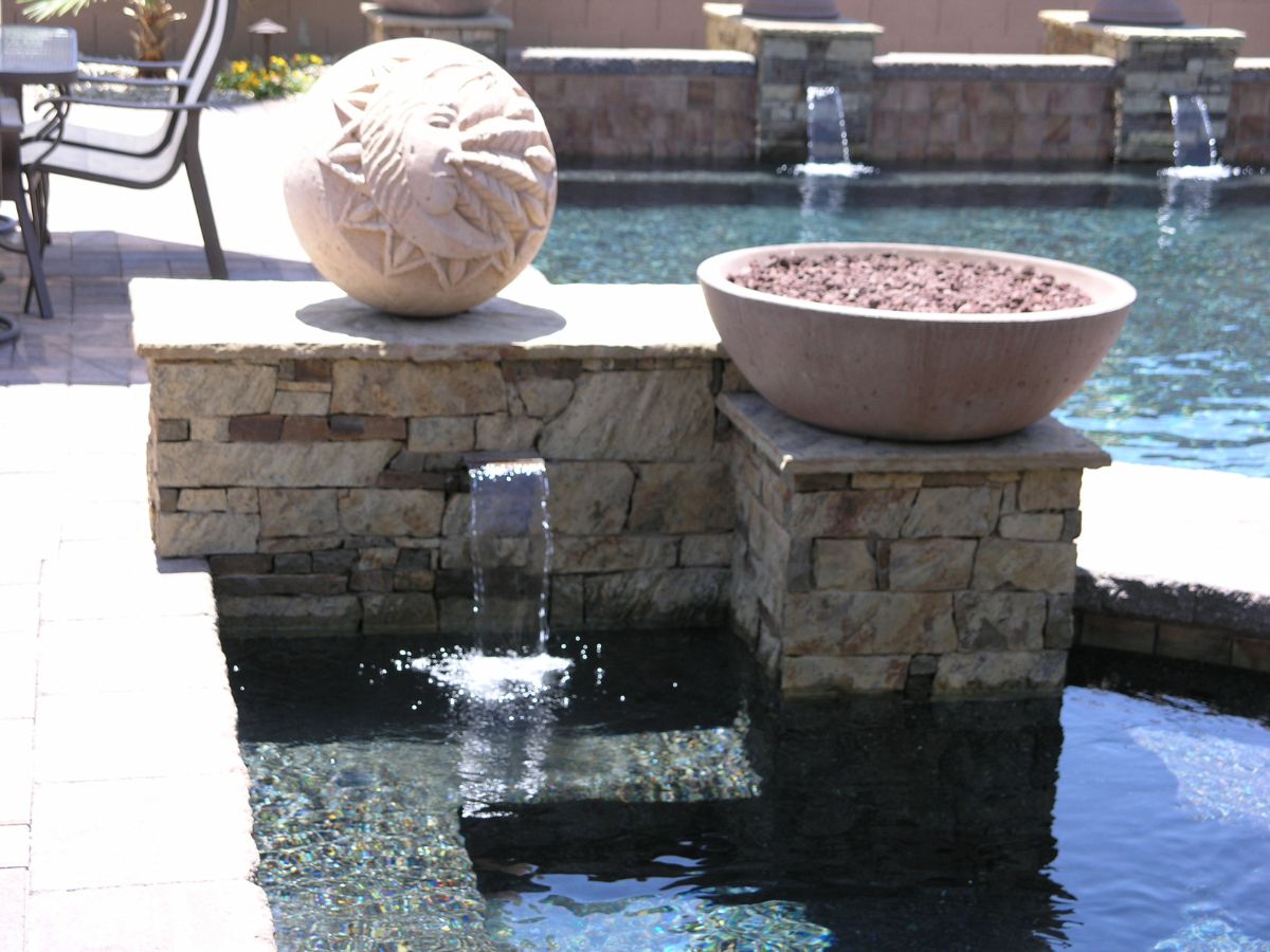 Backyard pool fountain with stone wall, decorative sun-etched orb and bowl planter spilling water into pool.