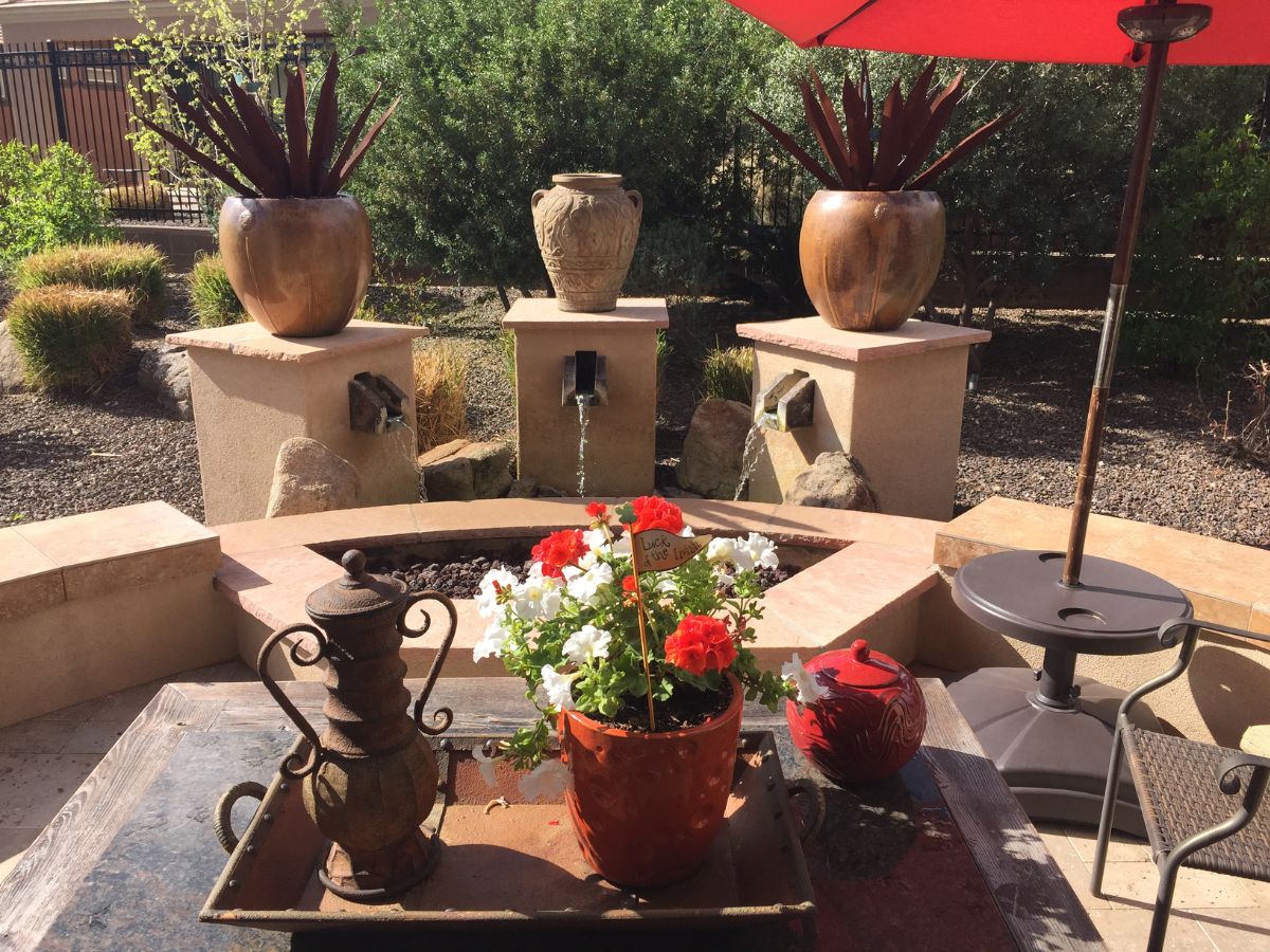 Sunny backyard patio with circular water fountain, terracotta pots, and red and white potted flowers under umbrella