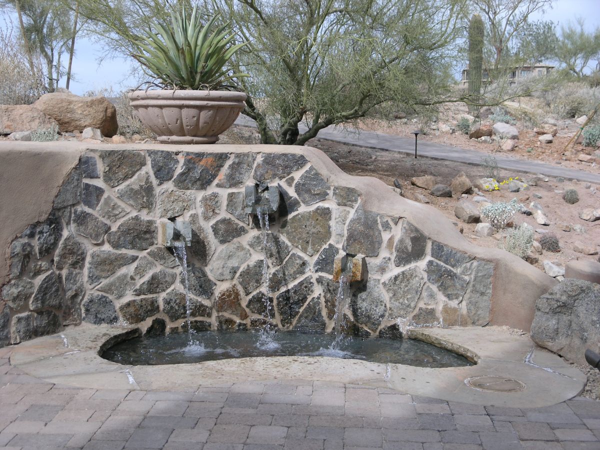 Desert garden stone wall fountain with three spouts flowing into a small pool, potted agave and xeriscape landscaping