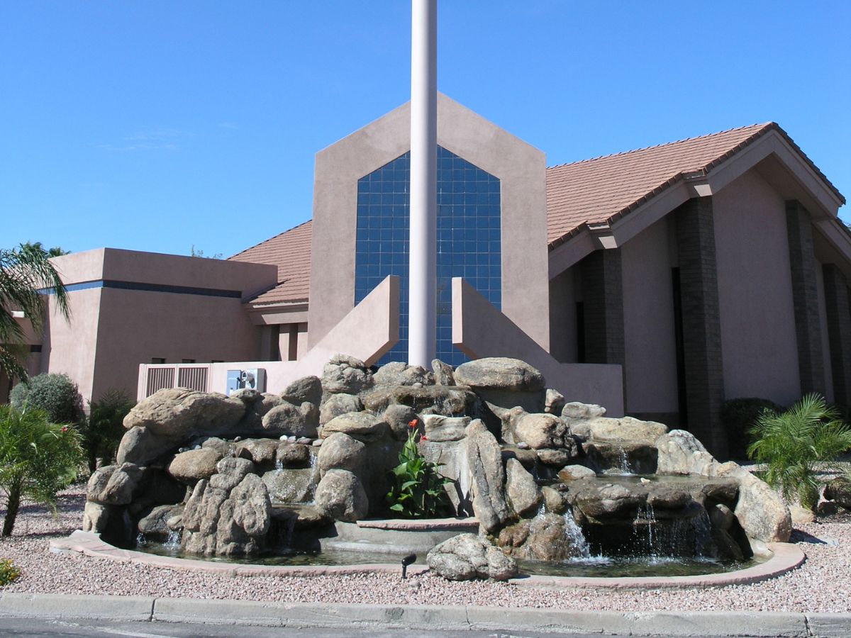 Exterior of modern church with large glass facade, flagpole and rock waterfall fountain in landscaped desert courtyard