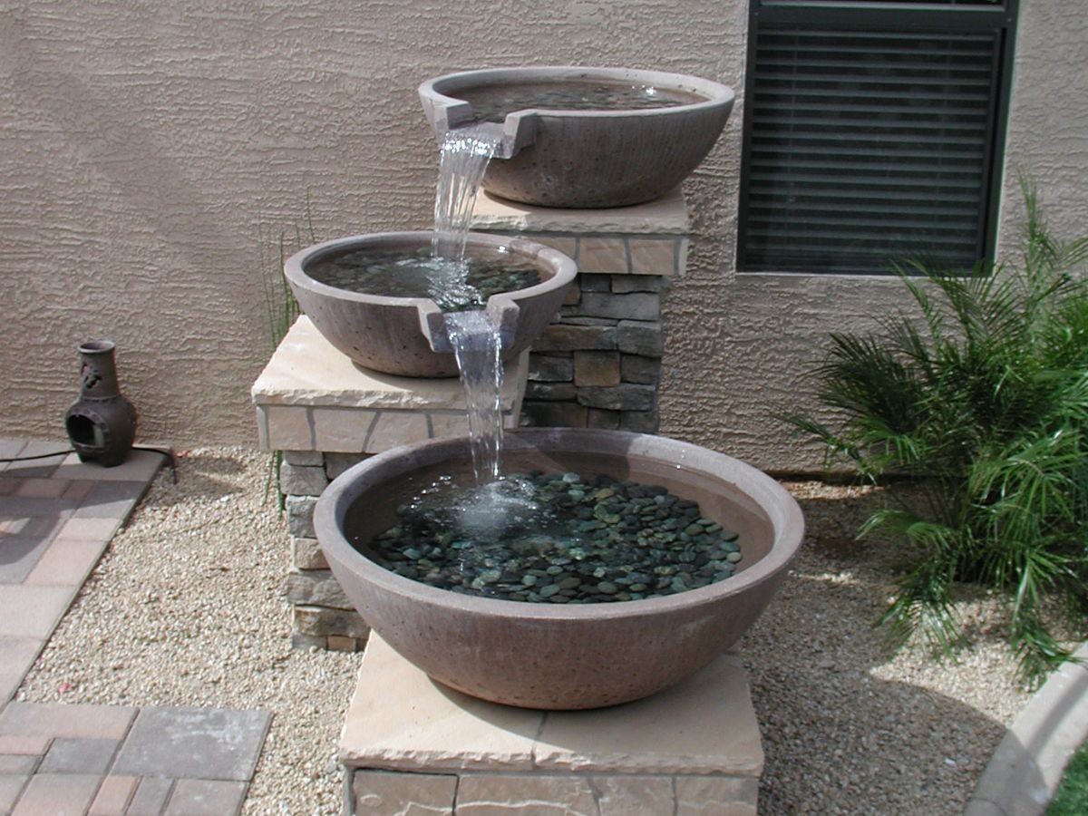 Outdoor three-tiered stone bowl water fountain with cascading water and dark pebbles on a patio garden