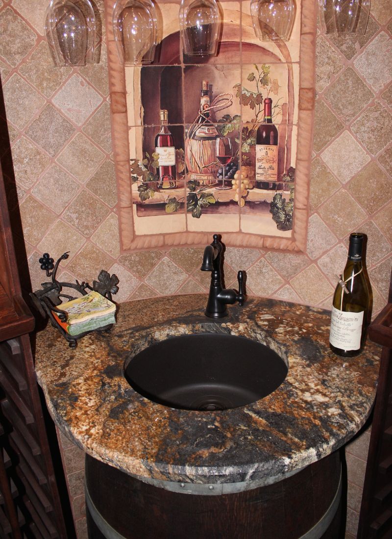 Wine-themed bathroom sink: black basin set in granite countertop on barrel base, wine tile mural & bottle
