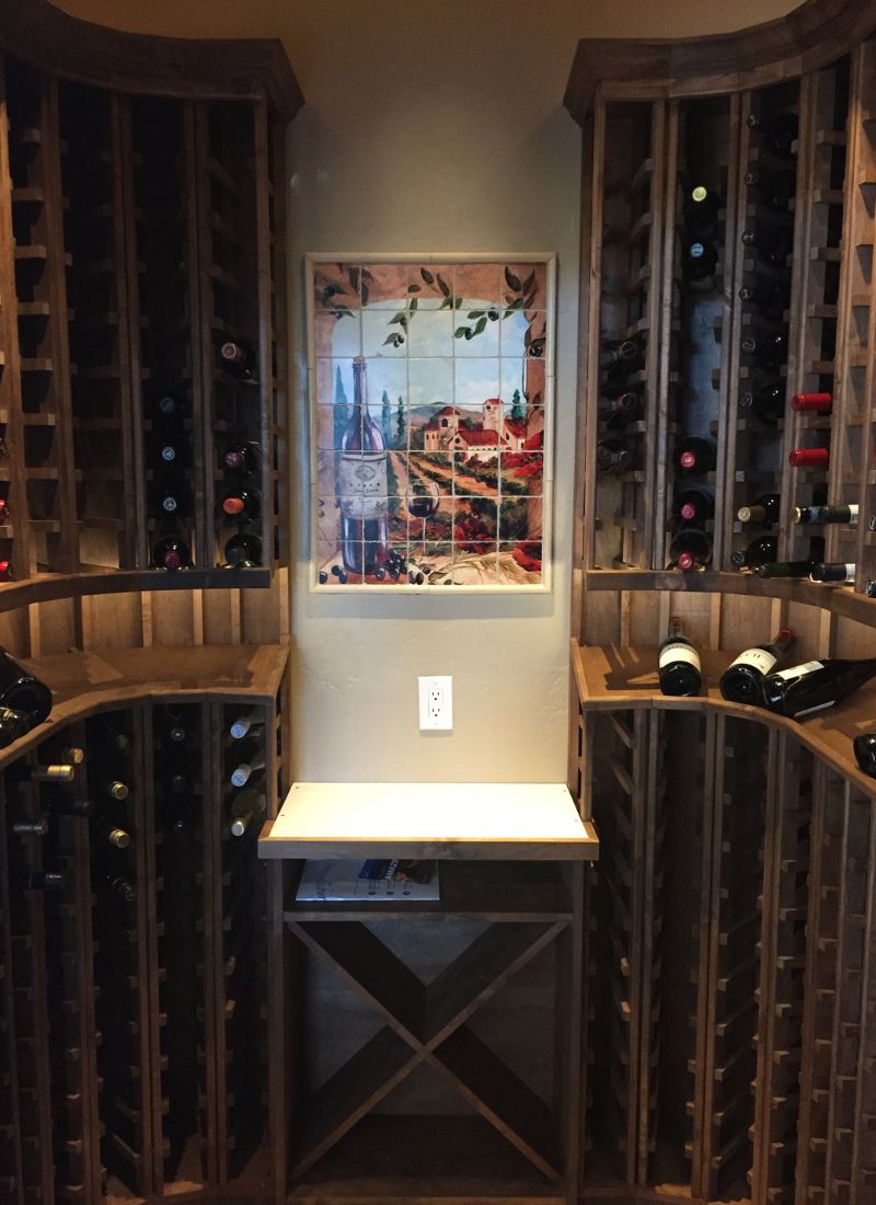 Wine cellar with curved wooden racks holding bottles, tiled vineyard mural and small tasting shelf.