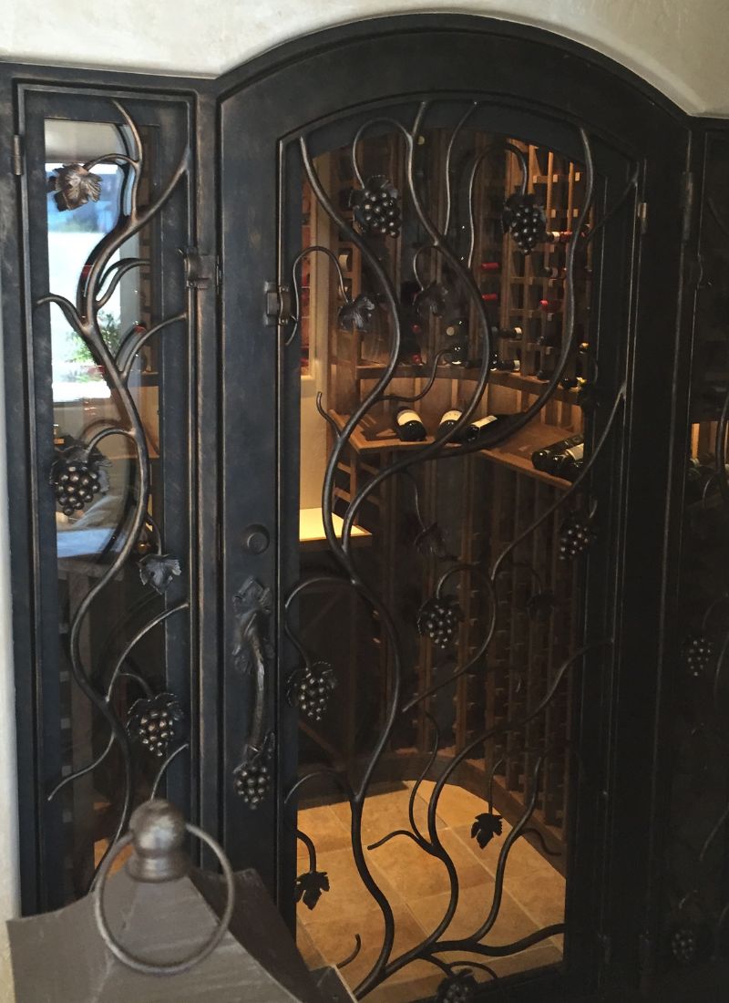 Ornate wrought-iron grapevine wine cellar door with glass panels revealing wooden wine racks and bottles inside